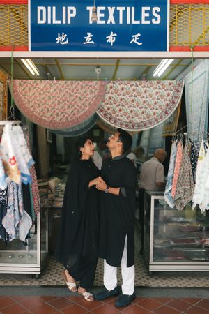 Couple in black traditional attire standing romantically outside Dilip Textiles fabric shop in Singapore.