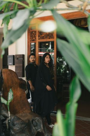 Two people dressed in black stand near a wooden door surrounded by tropical plants and sculptures.