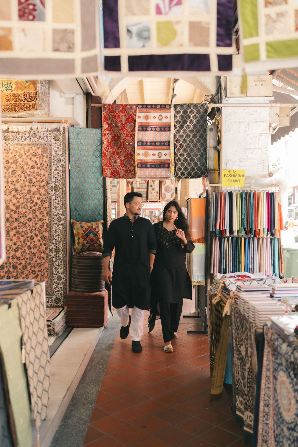 Couple in black traditional attire walking through a colorful fabric market with textiles displayed on both sides.
