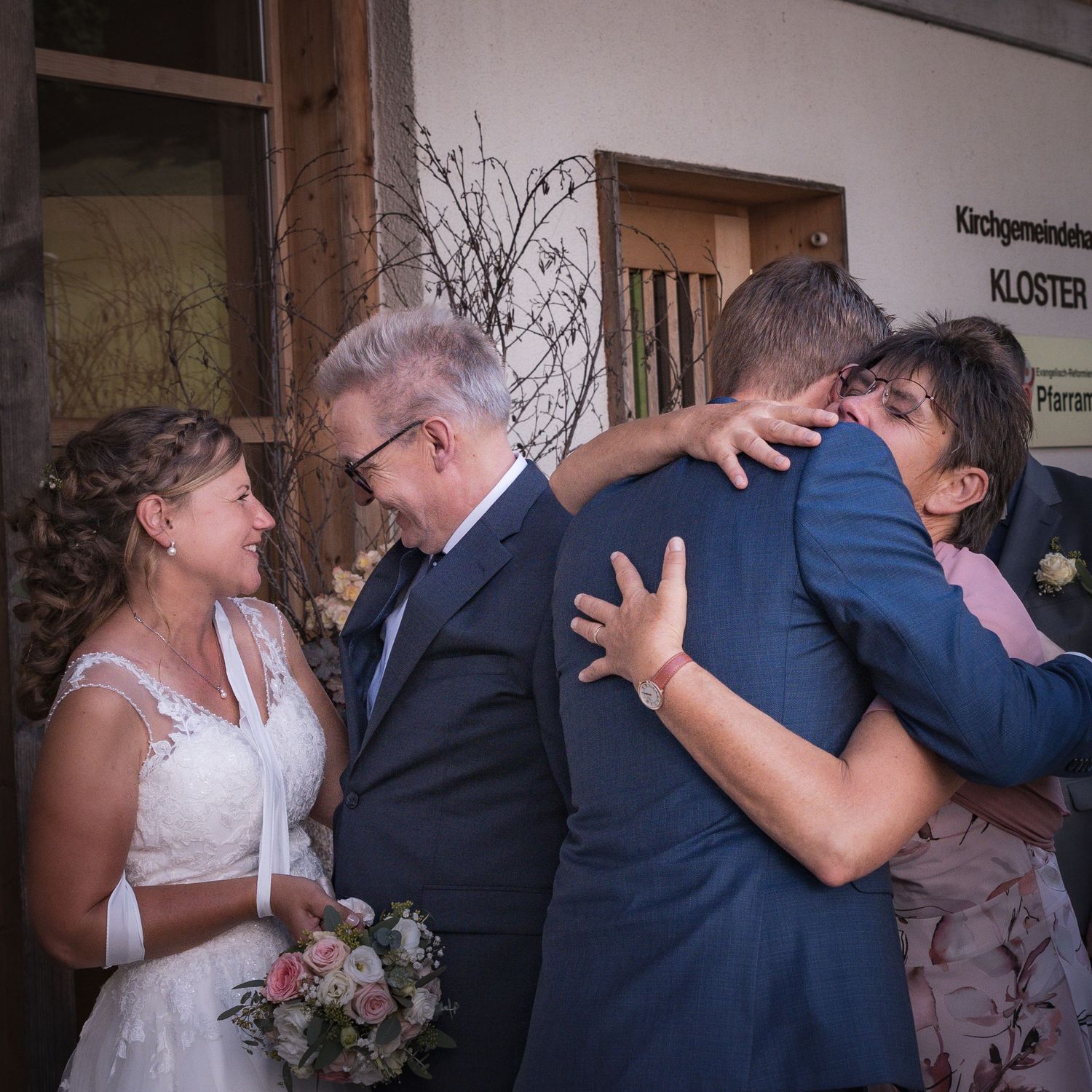 Bride in white dress and groom in blue suit embrace family outside a church building after wedding ceremony.