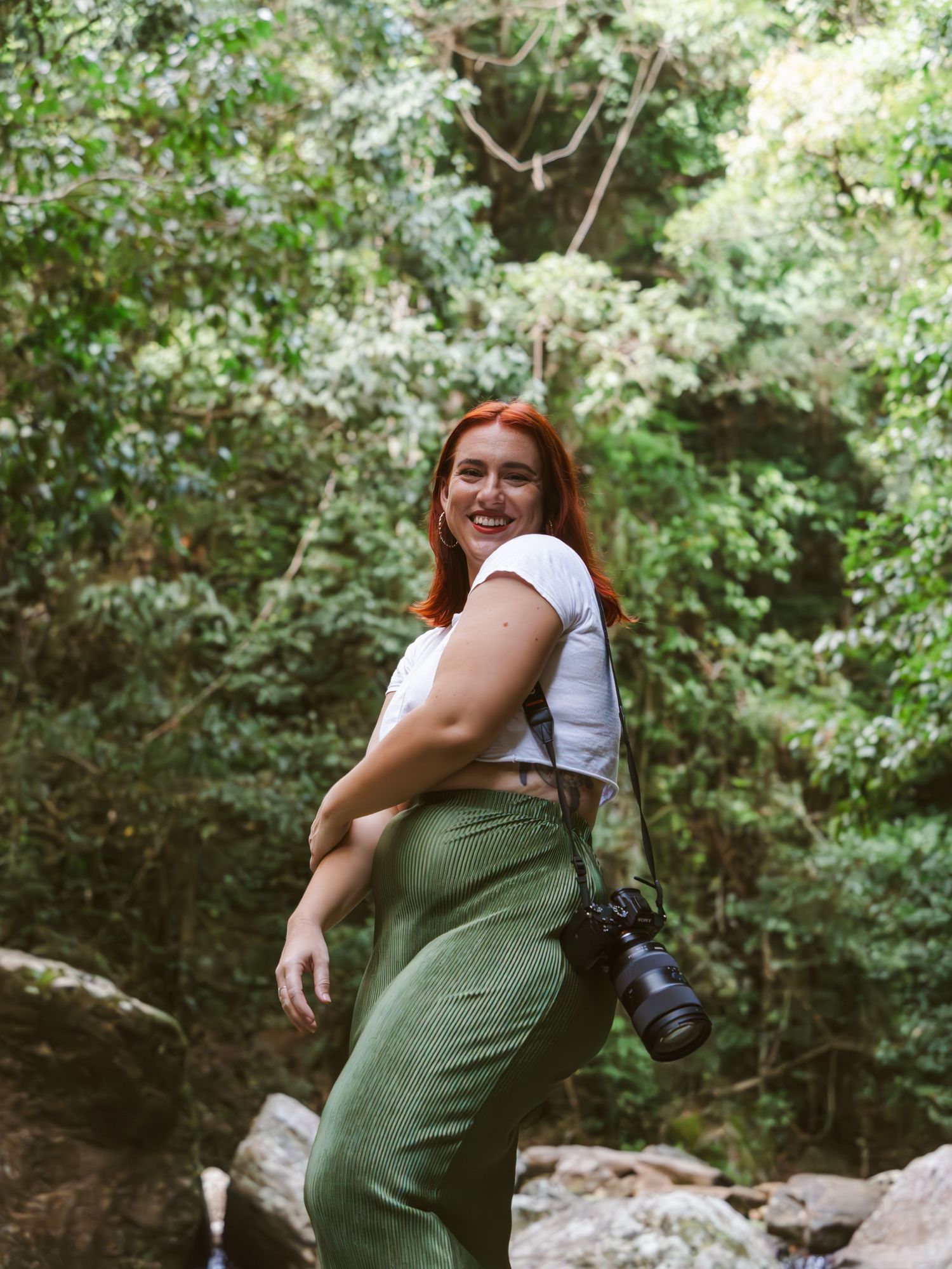 A smiling redhead woman with a camera explores a lush green forest, wearing a white crop top and green pants. Cairns boudoir photographer.