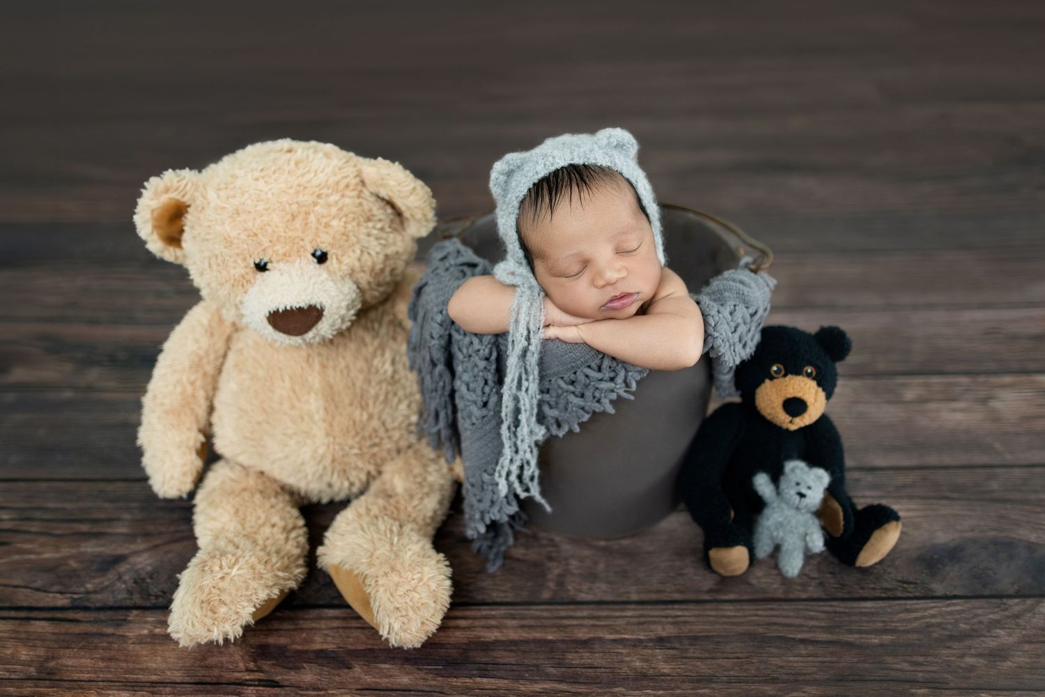 Newborn baby sleeping in gray bucket wearing blue bear bonnet, flanked by tan and black teddy bears on wood floor.