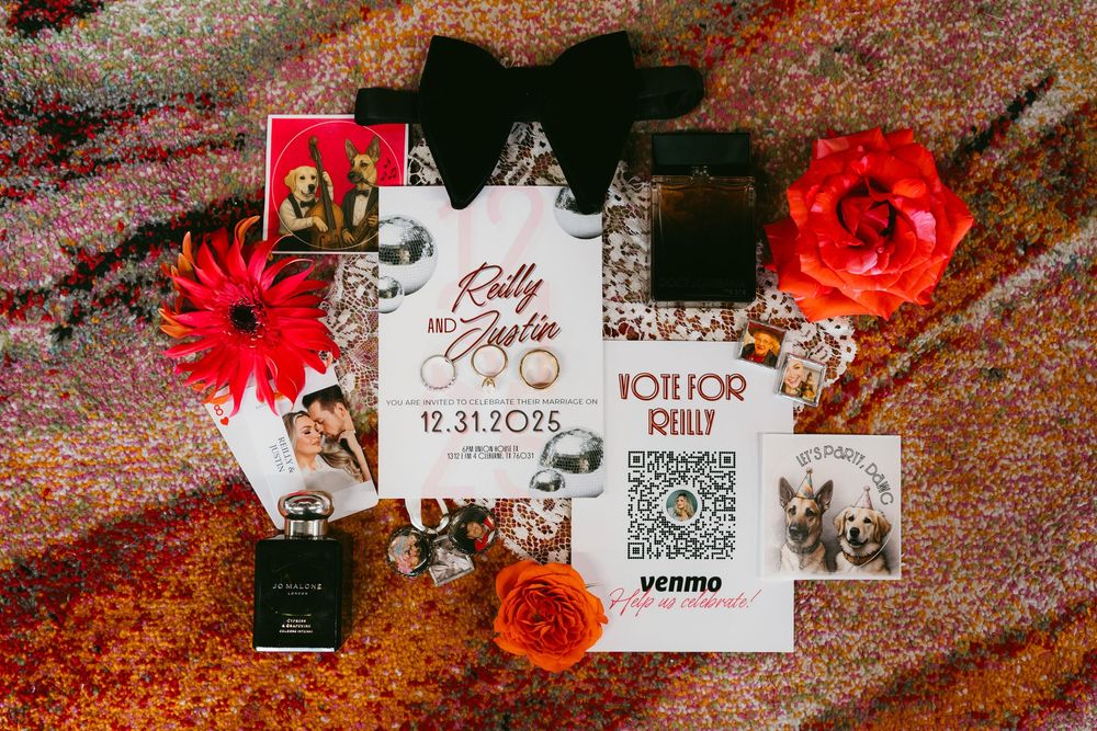 Elegant wedding stationery flat lay with red florals, perfume, rings, and a QR code Venmo card on a glittery surface.