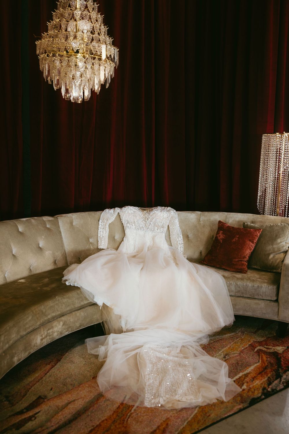 Elegant long-sleeve lace wedding gown displayed on a velvet curved sofa beneath a crystal chandelier with red curtains.