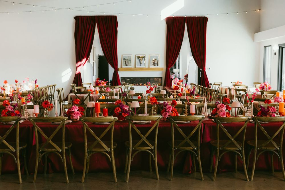 Elegant wedding reception with long tables, red linens, floral centerpieces, and burgundy drape curtains.
