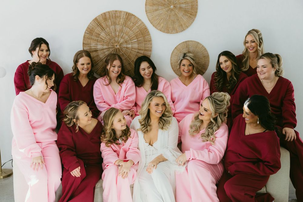 Bride in white robe surrounded by bridesmaids in pink and burgundy robes laughing together before wedding ceremony.