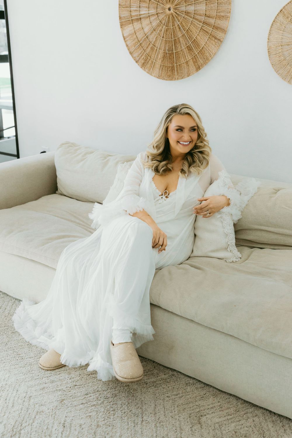 Smiling blonde bride in a white lace wedding gown sitting on a beige sofa wearing UGG slippers.