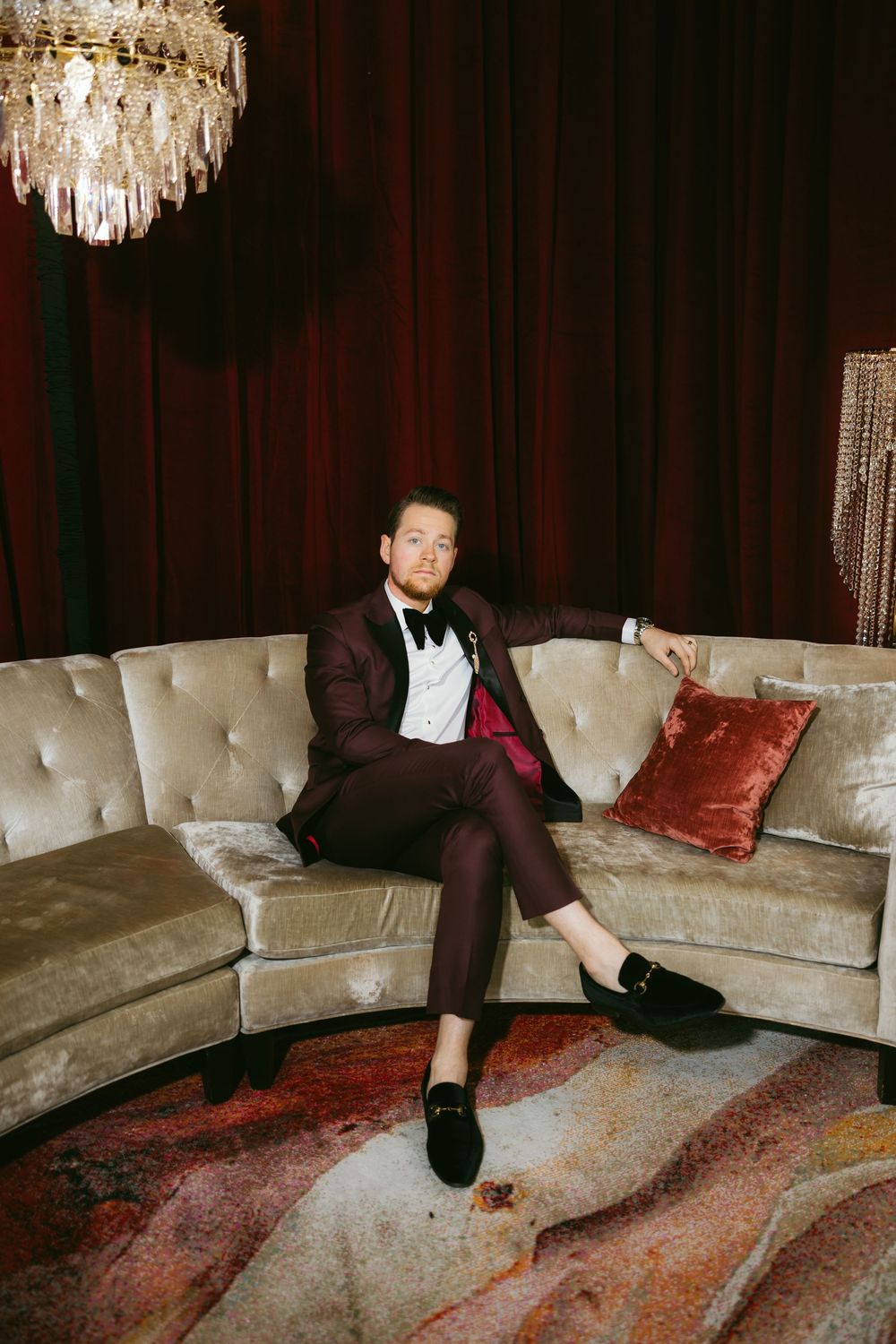 Man in burgundy tuxedo sitting on velvet sofa under crystal chandelier with red curtain backdrop.