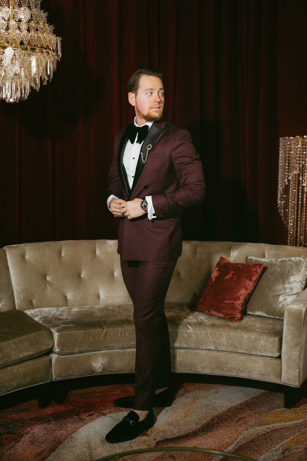 Groom in a burgundy tuxedo with black lapels standing beside a velvet sofa under a crystal chandelier.
