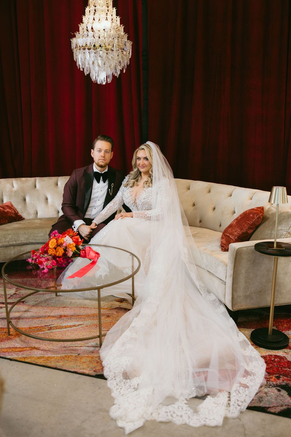 Bride and groom posing on a tufted sofa beneath a chandelier with red velvet drapes in background.