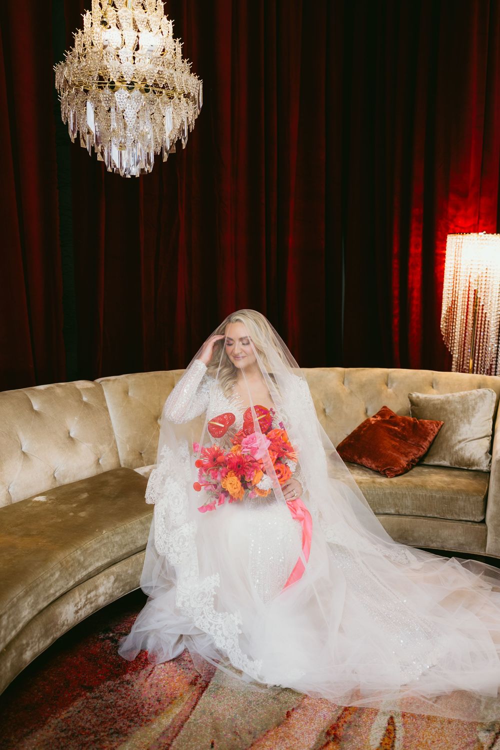 Bride in white gown and veil holds colorful bouquet on velvet sofa beneath crystal chandelier.