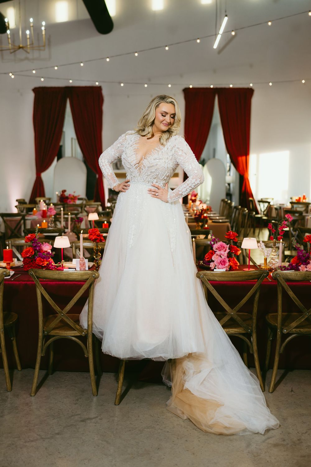 Bride in a long-sleeve lace wedding gown posing in front of a decorated red and gold reception table.