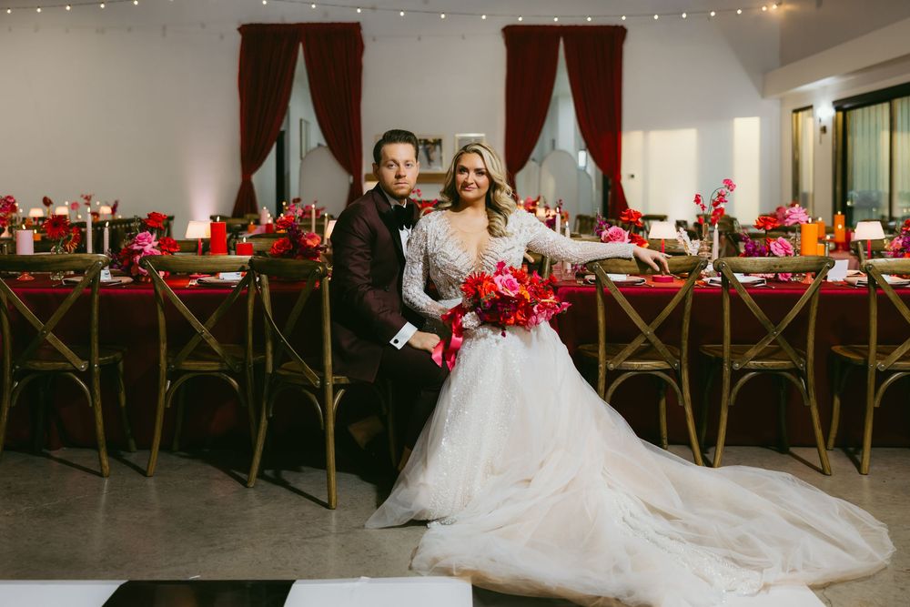 Bride and groom smiling together in front of an elegant red and pink floral wedding reception setup.