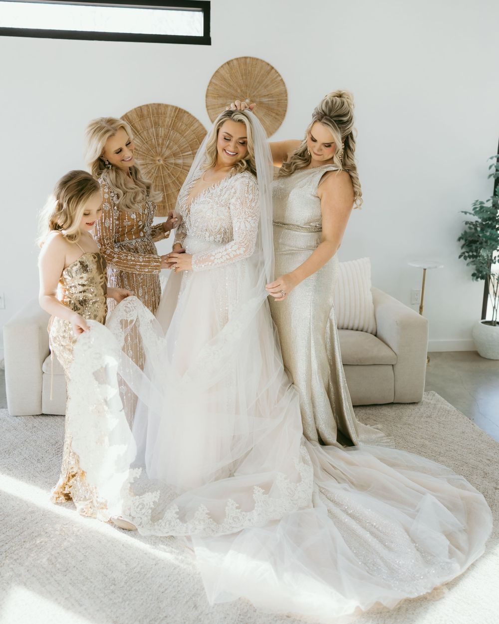 Bride in lace wedding gown with veil surrounded by bridesmaids in gold and champagne dresses in bright modern room.
