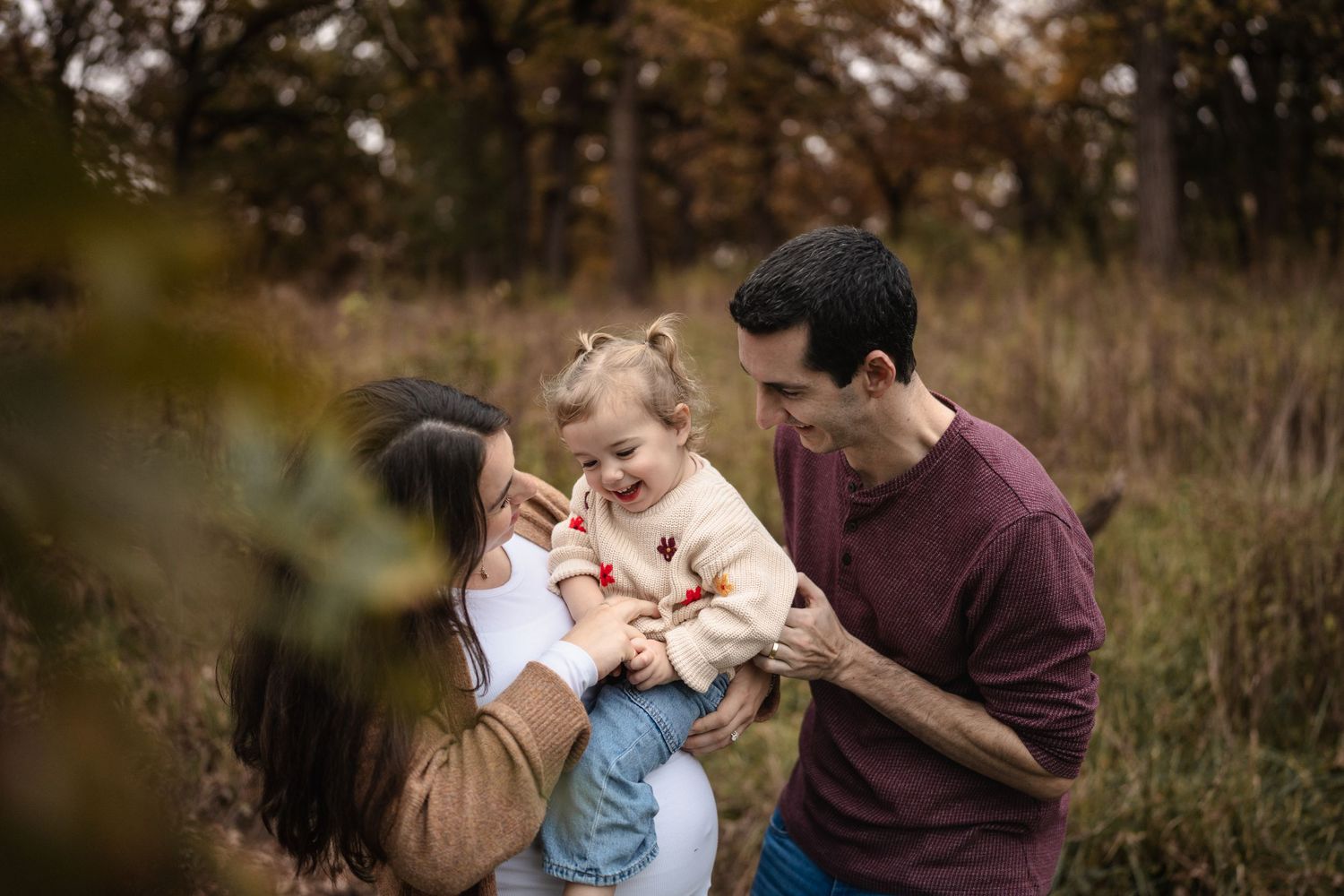 A happy young family with a toddler girl enjoys a warm autumn day in a golden meadow surrounded by fall foliage.
