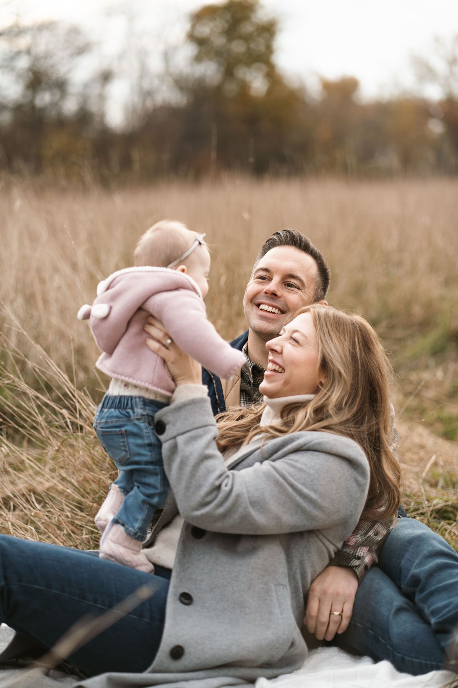 Happy couple sitting in autumn field holding smiling baby in pink hoodie during outdoor family photo session.