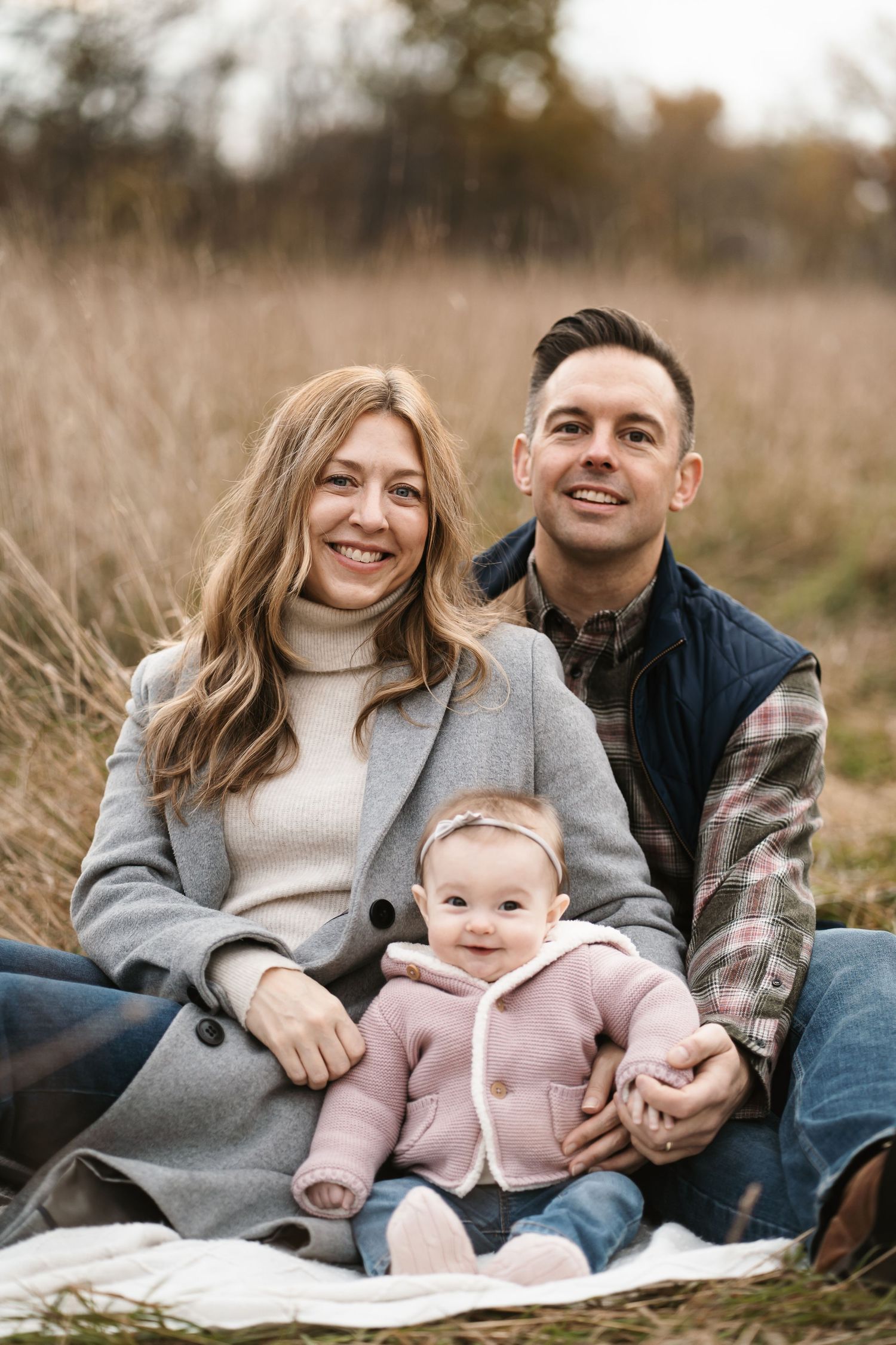 A happy family of three sits on a blanket in an autumn field, with parents smiling and a baby girl in a pink hoodie.