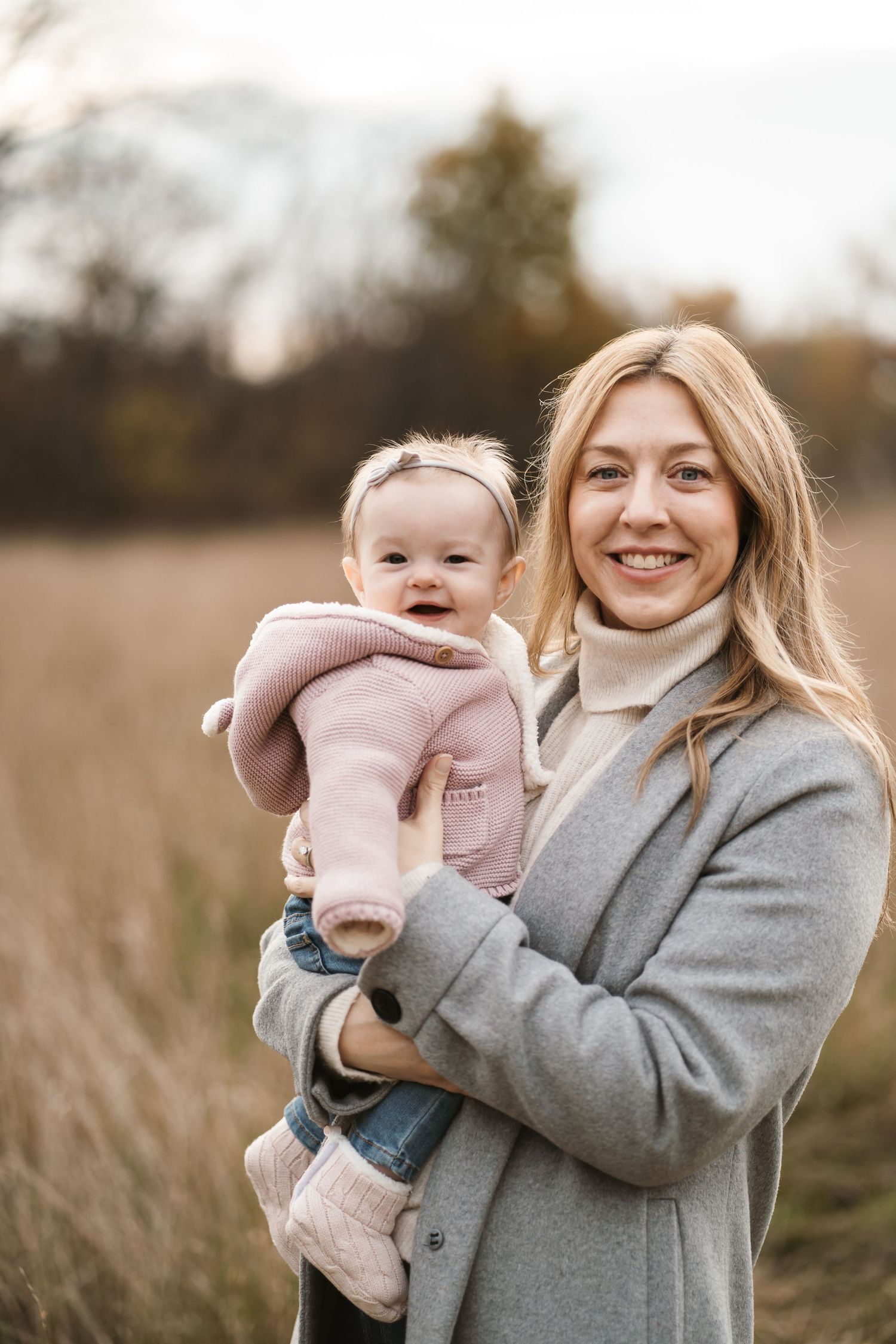 Smiling blonde woman in gray coat holding a happy baby in pink outfit in an autumn field.
