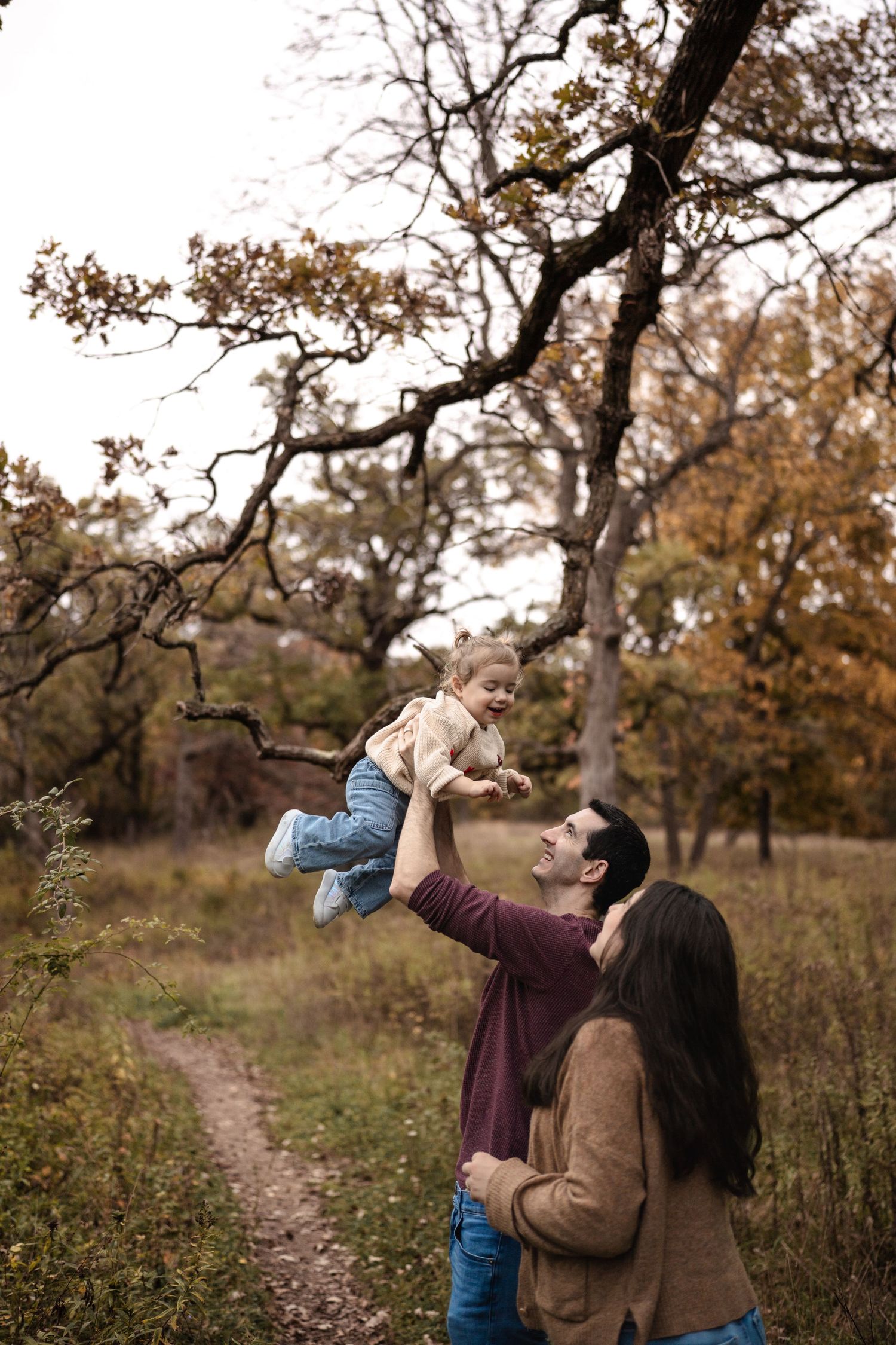 A happy family enjoys an autumn day outdoors, with a father lifting a smiling toddler high in the air among fall foliage.