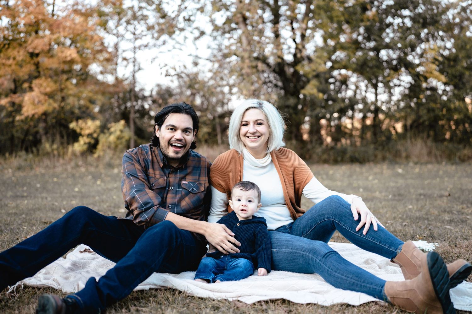 A smiling family of three sits on a blanket in an autumn field, with colorful fall foliage in the background.