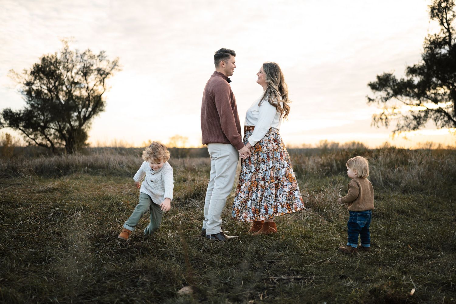 Family of four poses at golden hour in a field, parents holding hands while two toddlers play around them.