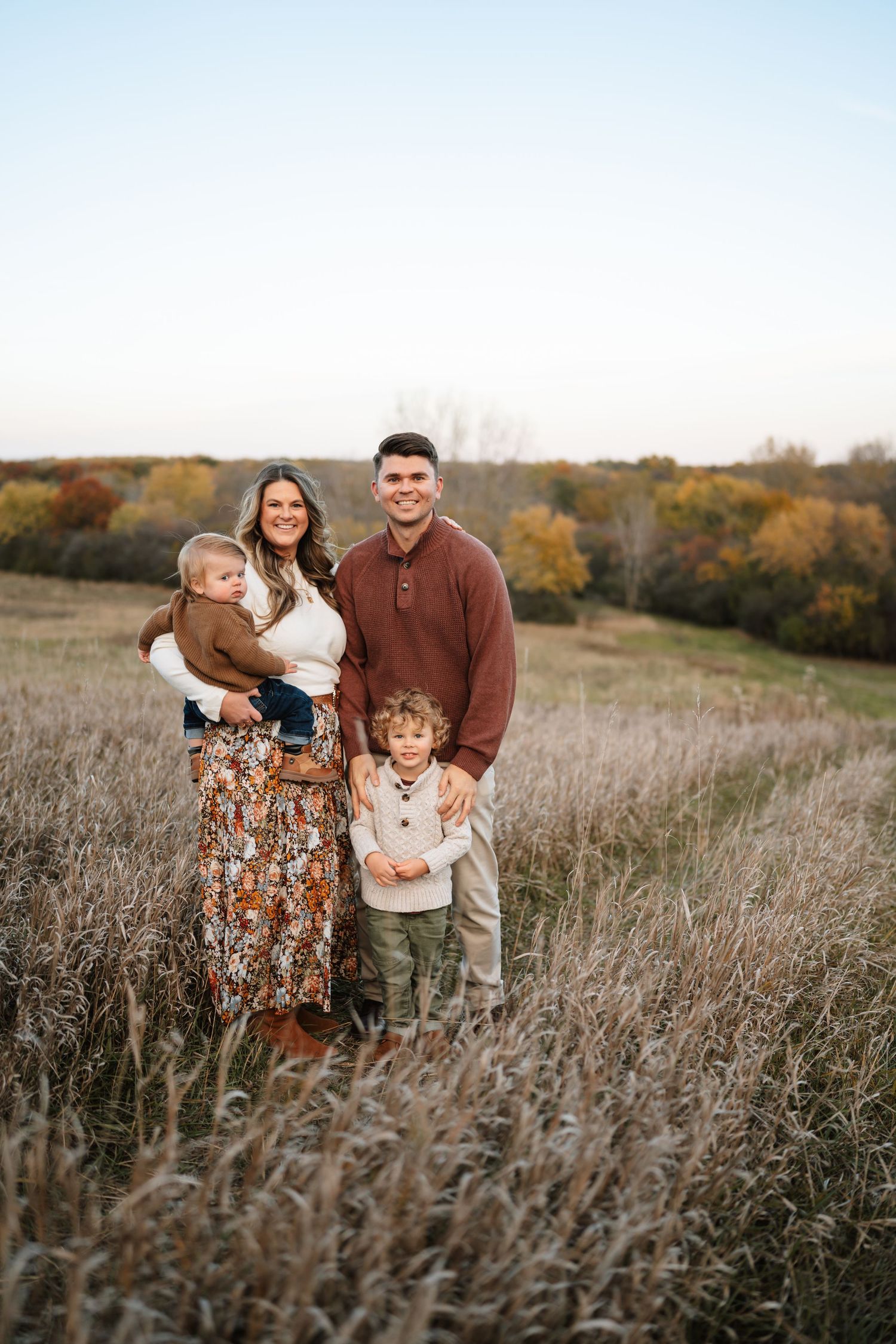 Family of four posing in autumn field with tall grass and colorful fall foliage in background.