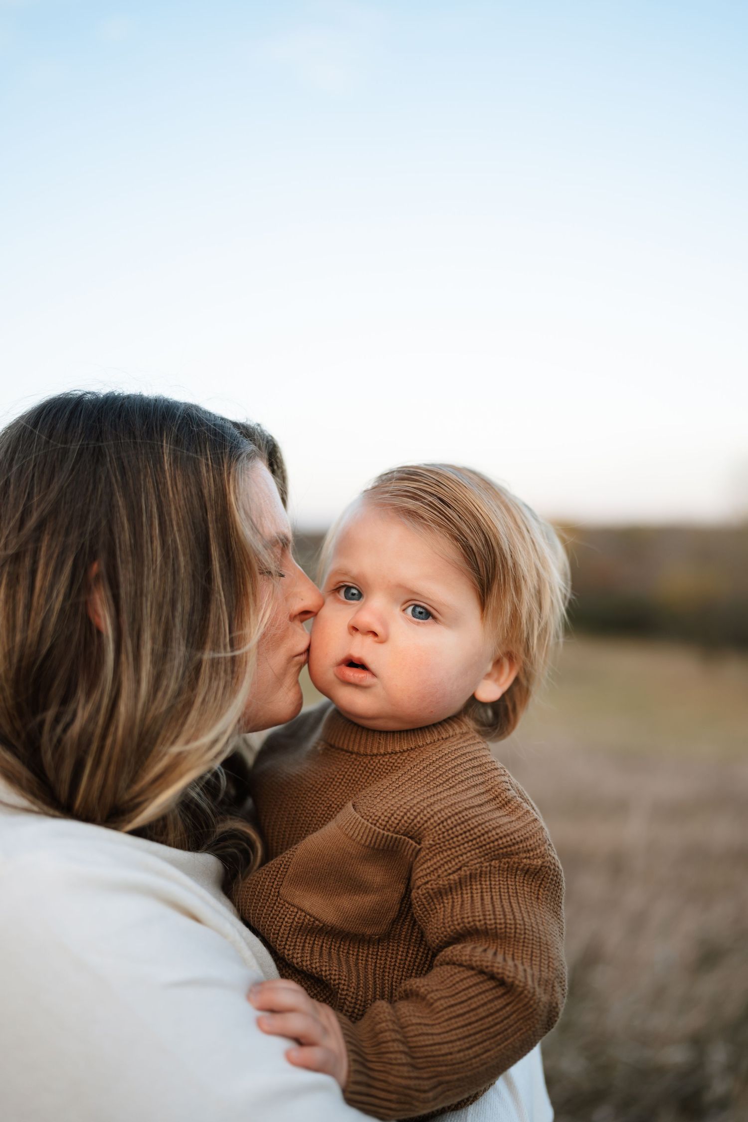 Mother kissing cute blonde toddler in brown sweater outdoors in natural light.