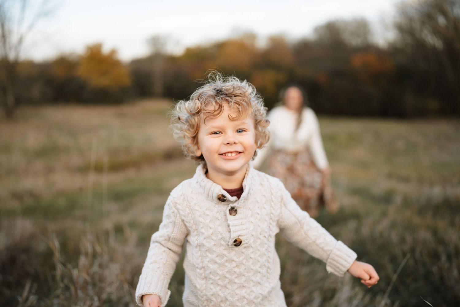 Curly-haired toddler boy smiling in autumn field wearing cream knit sweater with mother in background.