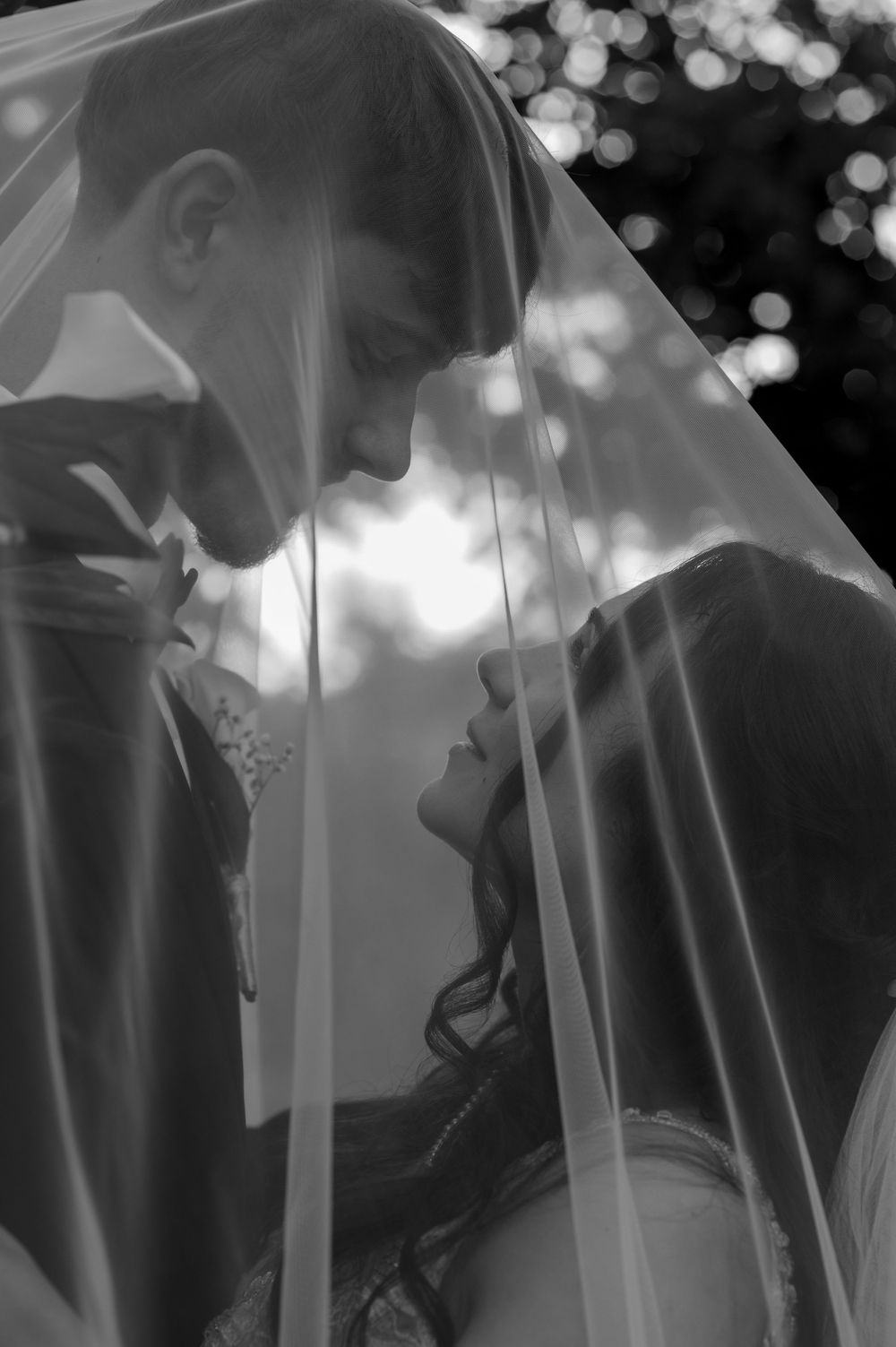 Black and white photo of bride and groom sharing an intimate moment under a flowing wedding veil.