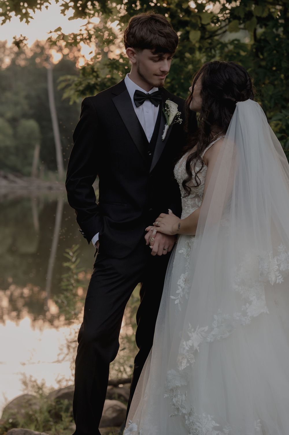Bride and groom holding hands by a reflective pond at golden hour in formal wedding attire.