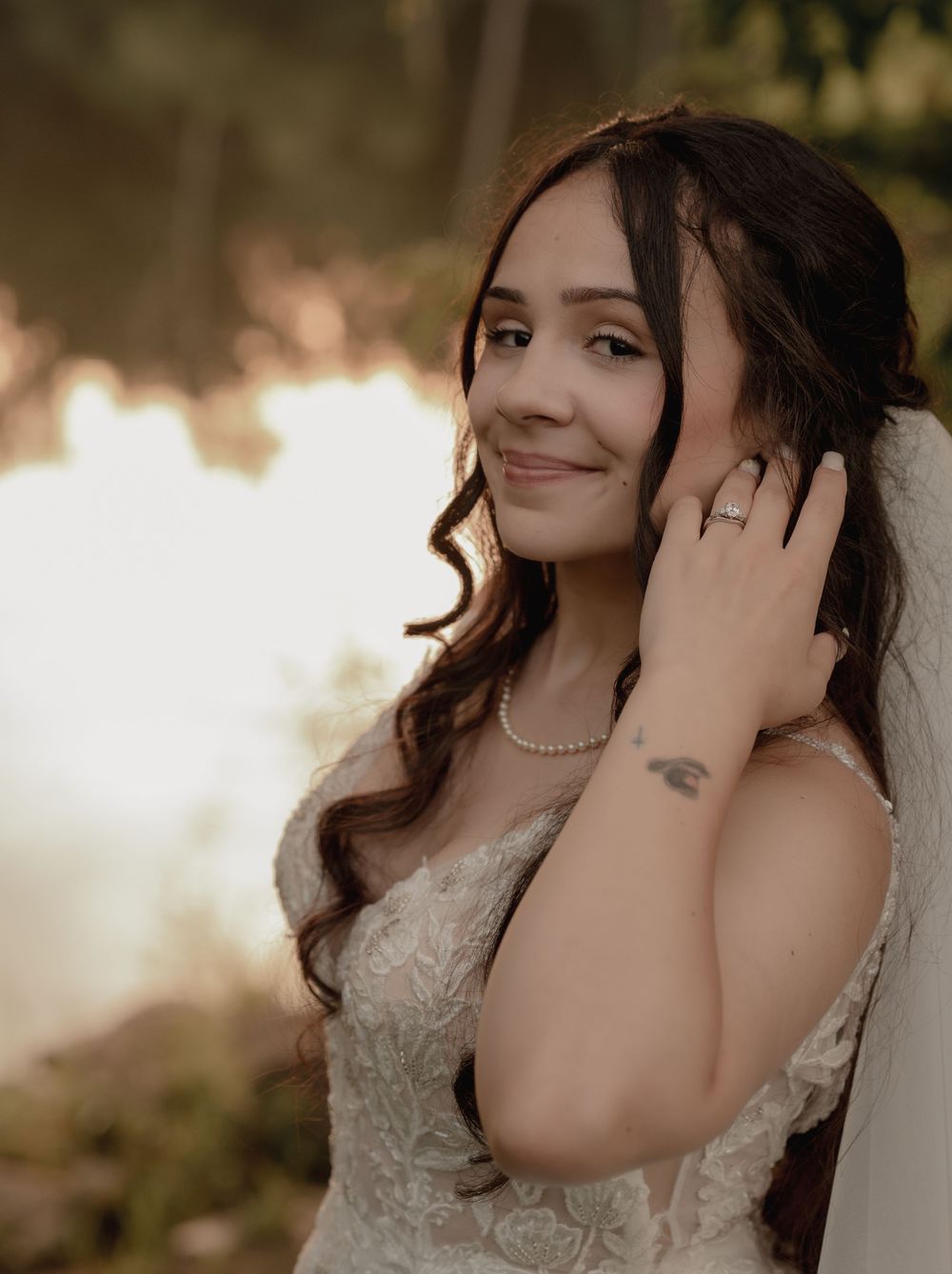 Bride in lace wedding gown poses gracefully outdoors with golden bokeh background, wearing pearl necklace and ring.