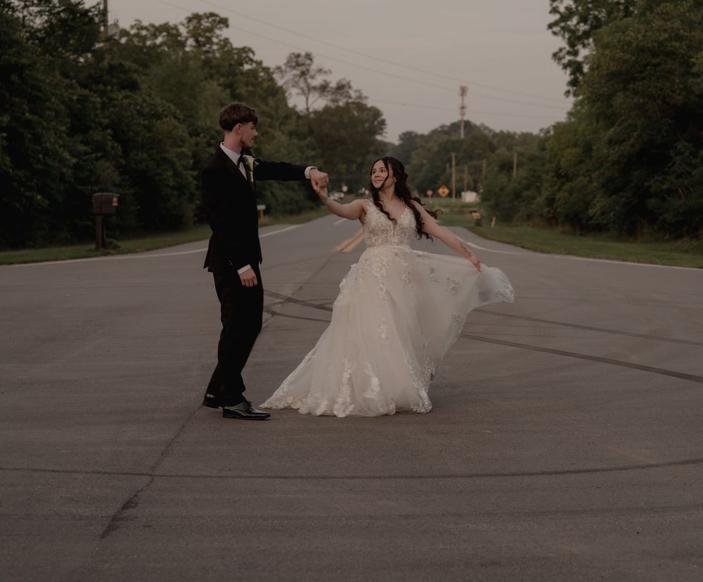 Bride in flowing white gown dancing with groom in black suit on open road surrounded by lush trees.