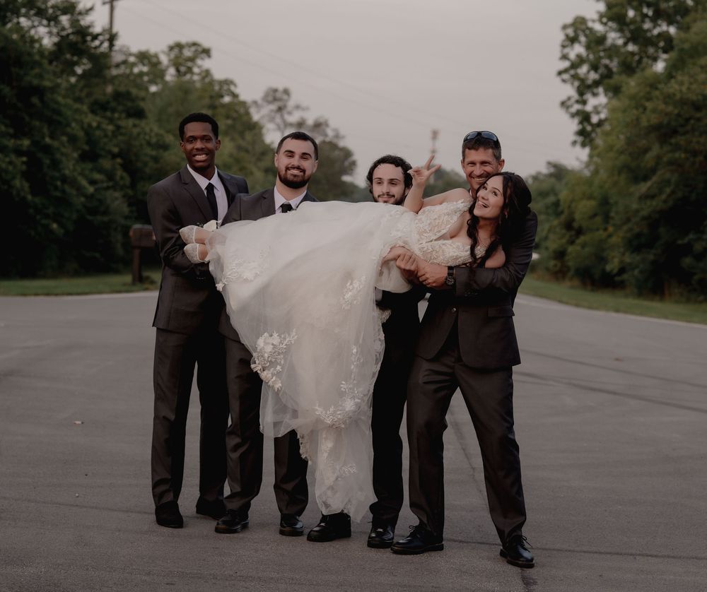 Groomsmen lifting bride in white wedding gown outdoors on pavement, celebrating with joy.