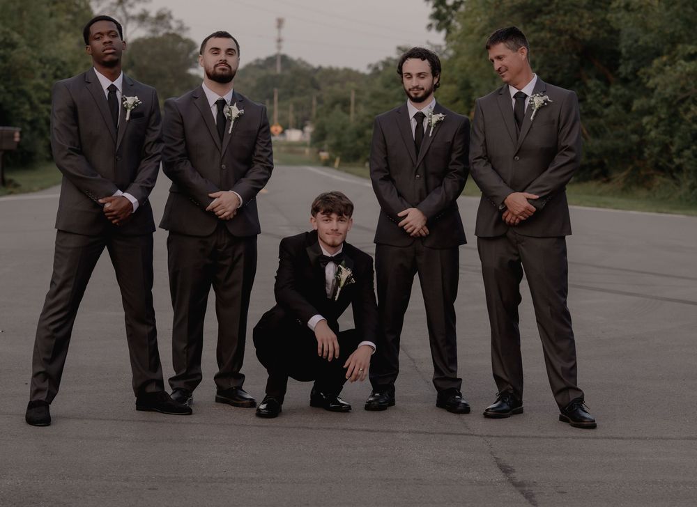 Five groomsmen in gray suits posing on open road with one crouching in front center.