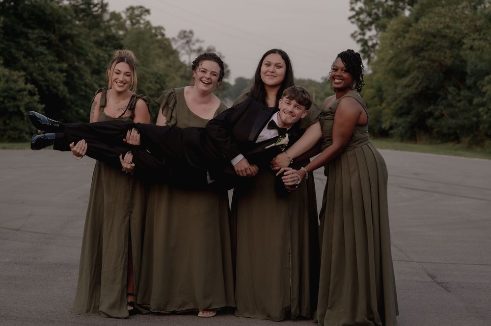 Four bridesmaids in olive green gowns playfully carry groom in black tuxedo on outdoor pavement.