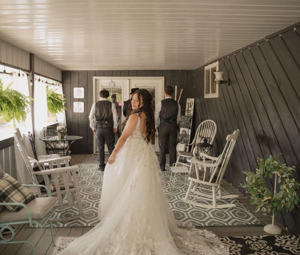 Bride in lace wedding gown stands in rustic gray-paneled venue with rocking chairs and greenery.