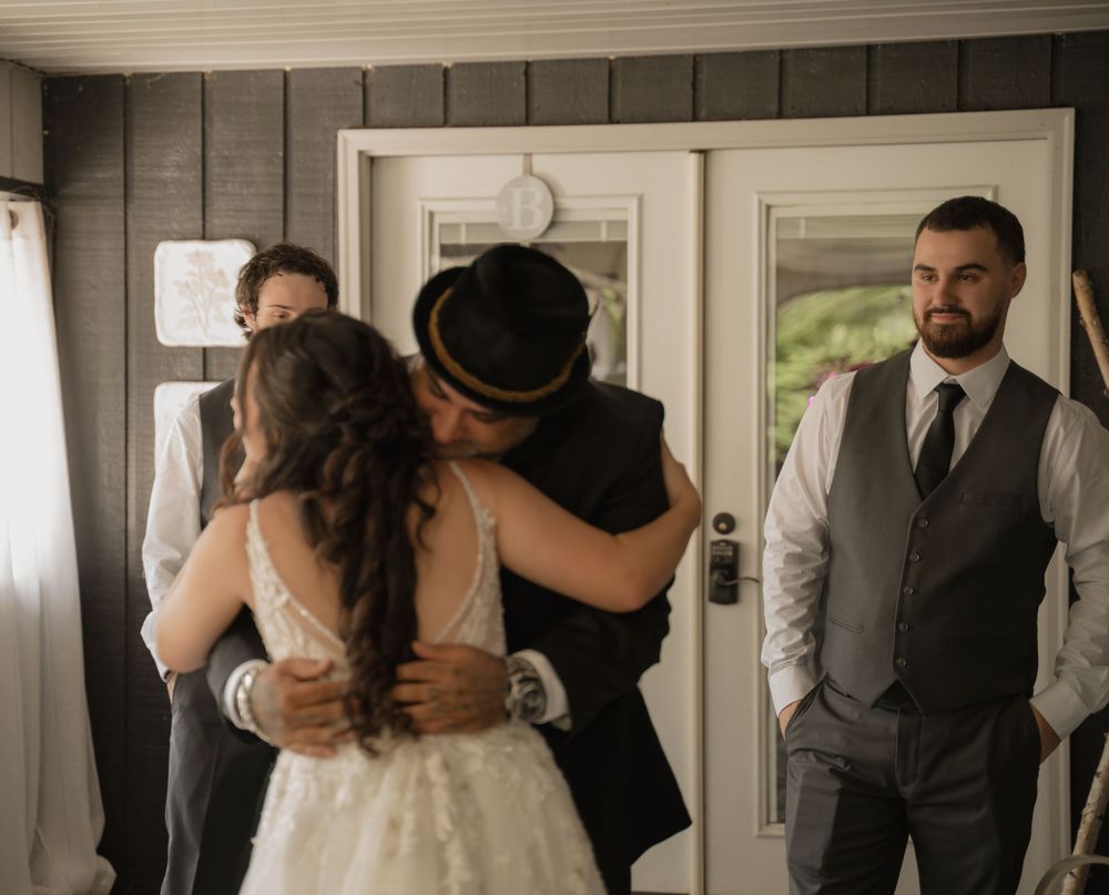 Emotional wedding moment as bride and guest embrace warmly indoors while groom watches nearby.