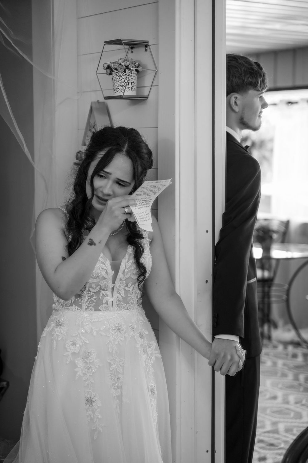 Black and white photo of emotional bride reading vow letter while holding grooms hand at door.