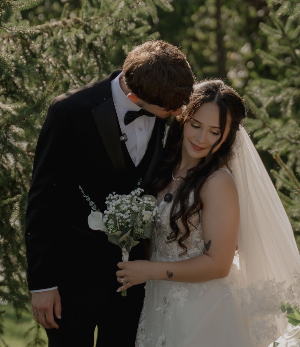 Groom in black tuxedo kissing bride holding white bouquet near evergreen trees.