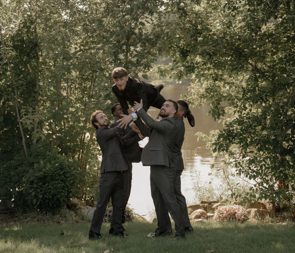 Three groomsmen playfully lifting one another in a lush green outdoor wedding photo.