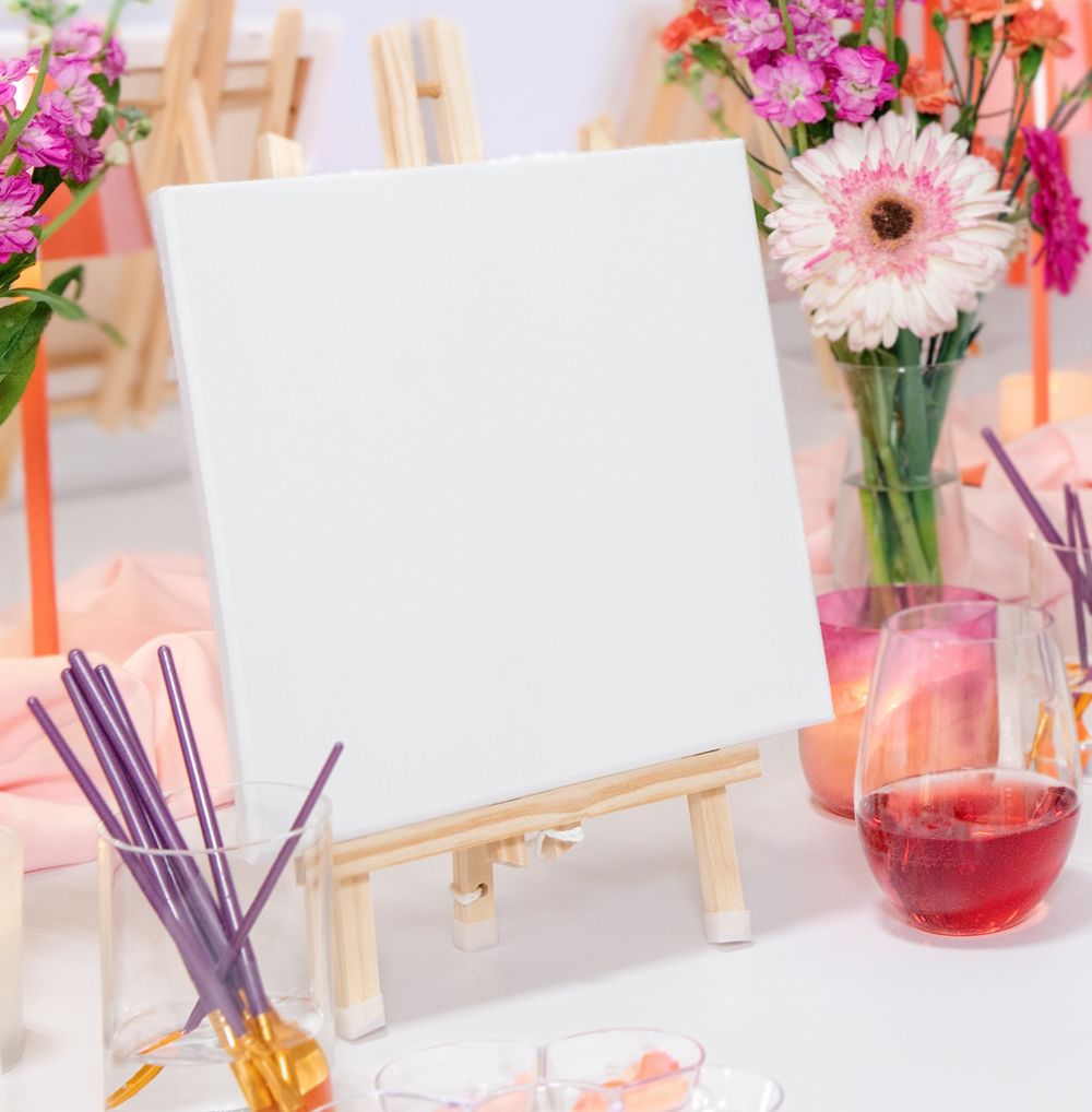 Blank white canvas on a wooden easel surrounded by colorful flowers, paint brushes, and a glass of red wine.