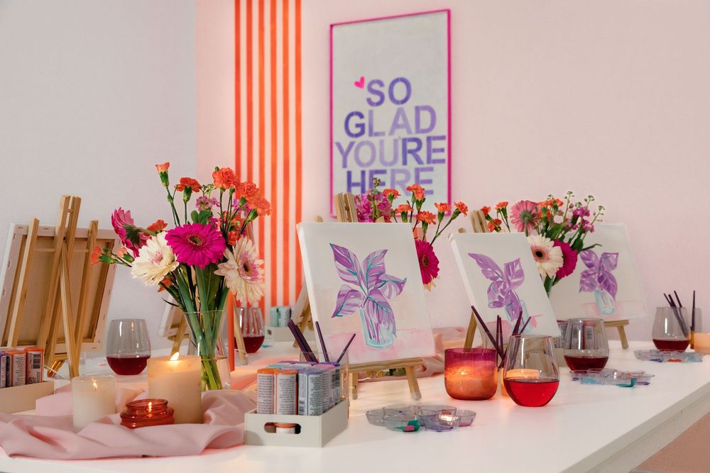 Art class setup with painted canvases on easels, fresh flowers, wine glasses, and a colorful 'So Glad You're Here' sign.