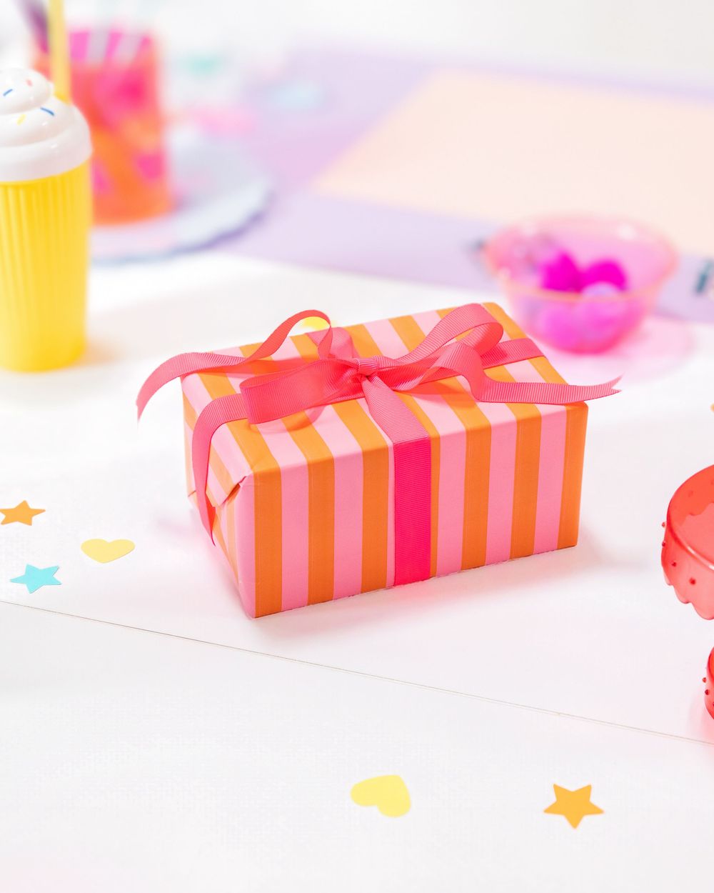 Pink and orange striped gift box with hot pink ribbon bow on white table with colorful confetti stars and hearts.