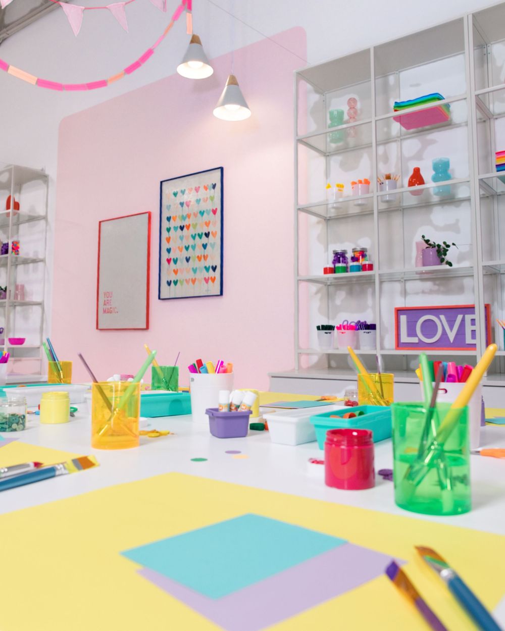Bright colorful art studio with pink walls, white shelves, craft supplies, and a LOVE sign on display.