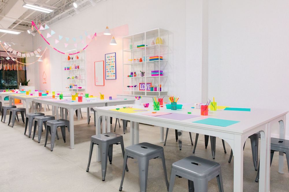 Bright art studio with white tables, colorful supplies, and metal stools in a cheerful kids classroom setting.