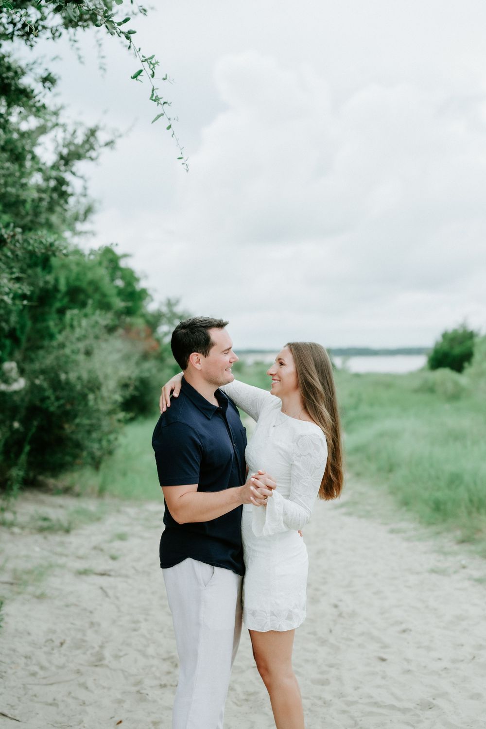 Couple embracing and smiling at each other on a sandy beach path surrounded by lush greenery.