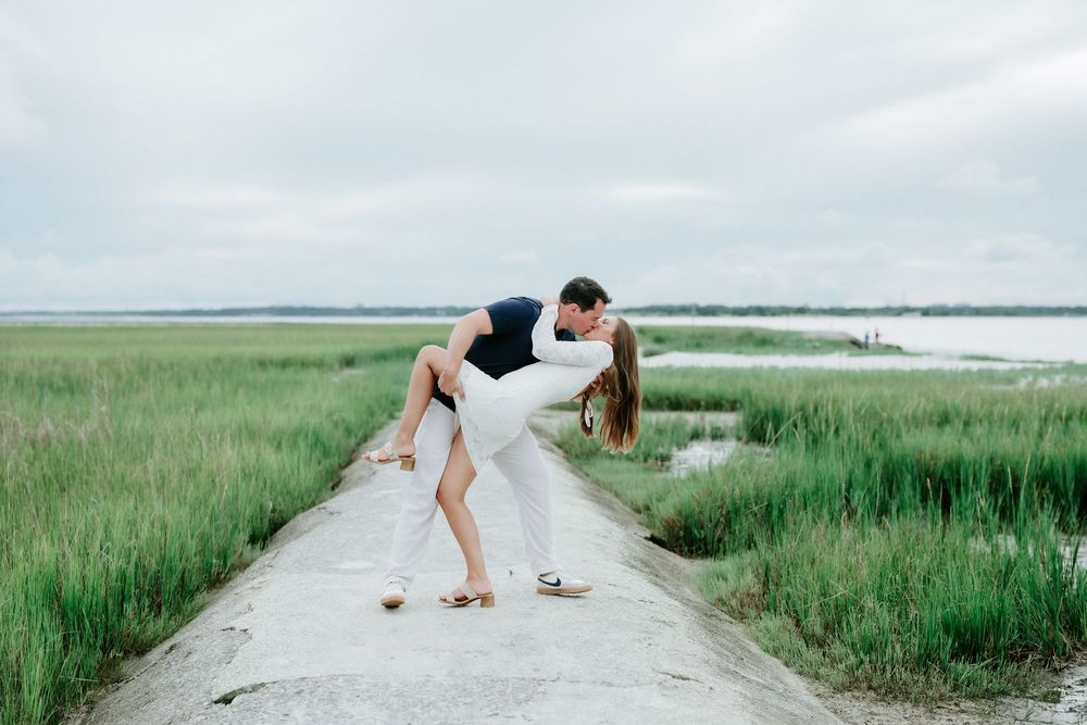 Couple sharing a romantic dip kiss on a coastal path surrounded by lush green marsh grass and calm waters.