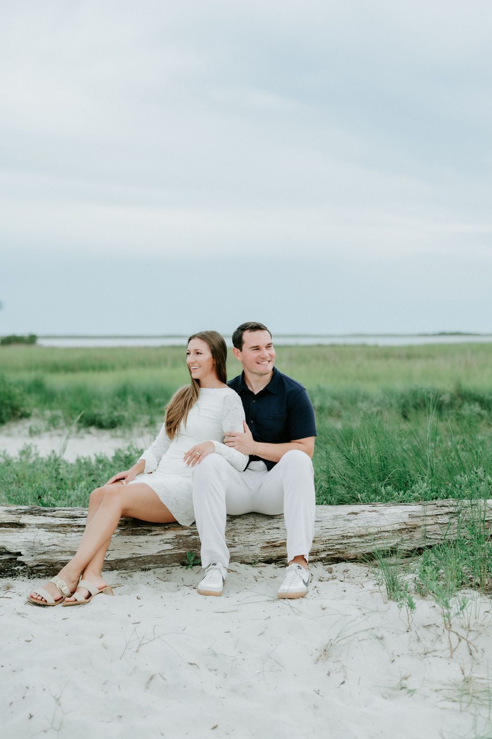 A couple sits together on driftwood at a sandy beach, surrounded by green marsh grass under a cloudy sky.