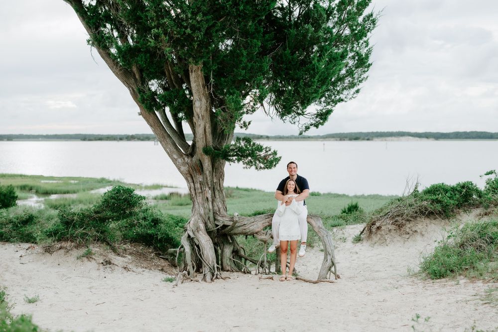 Couple embracing under a large tree on sandy beach with serene water and green landscape in background.
