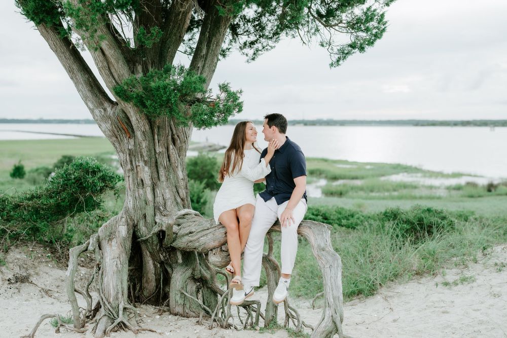 Couple sharing a romantic kiss while sitting on tree roots near a sandy beach with lush greenery and water in the background.
